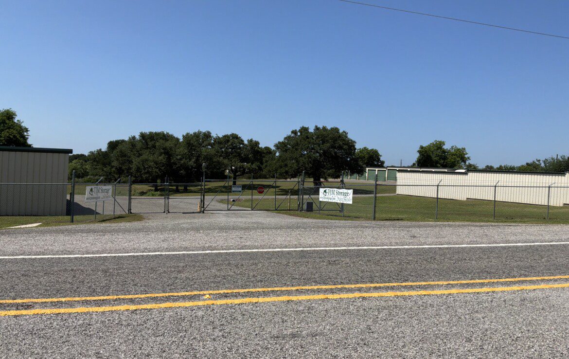 Roadside fence with trees and buildings visible.
